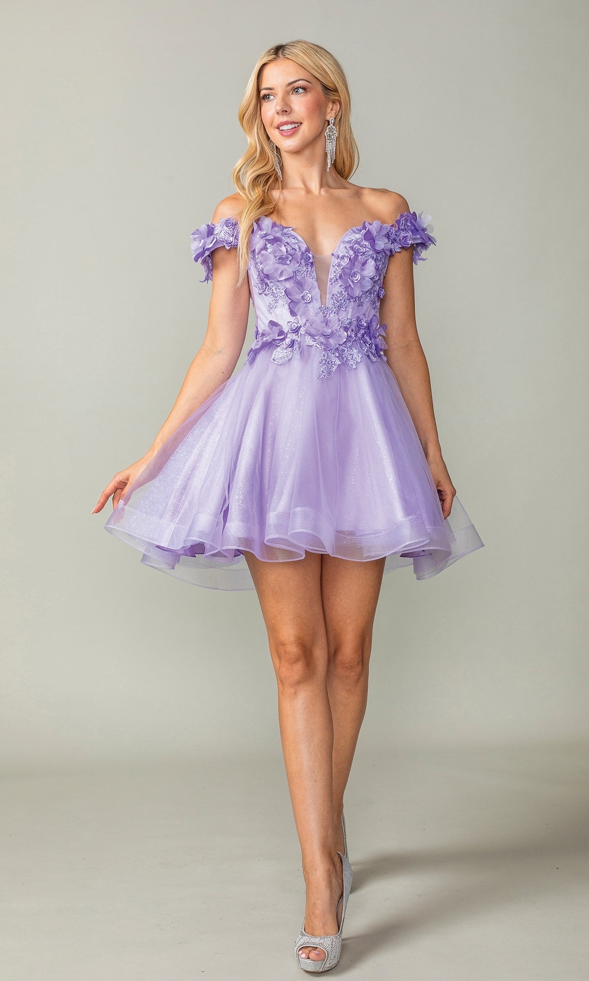 A woman models the Off-the-Shoulder Short Babydoll Hoco Dress 3344, featuring floral appliques and sheer details, as she stands against a plain light background.